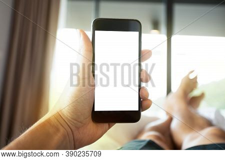 Close Up Image Of Man's Hand Holding Mobile Phone With Blank White Screen In The Bed Room. Man Layin