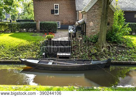 A Wooden Black Boat Is Moored In A Canal By The Shore In Front Of The House, The Broadside And Rudde