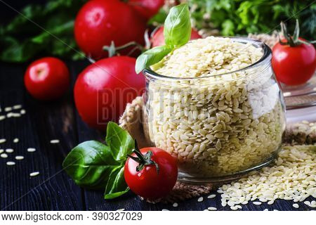 Italian Uncooked Pasta Riso Or Orzo In A Glass Jar, Tomatoes And Green Basil, Black Background, Sele