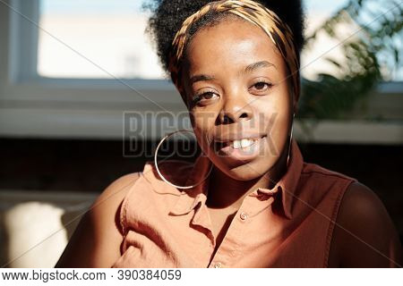 Young cheerful restful woman of African ethnicity looking at you with toothy smile while sitting by window in home environemnt