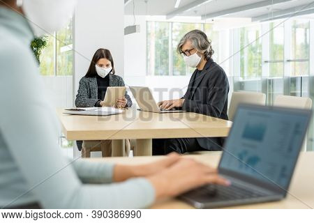 Two busy colleagues in smart casualwear and protective masks sitting by table and using mobile gagdets while networking in large office