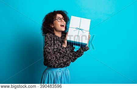 Surprised Woman Is Holding A Present And Posing With Glasses On A Blue Studio Wall Wearing A Dress A