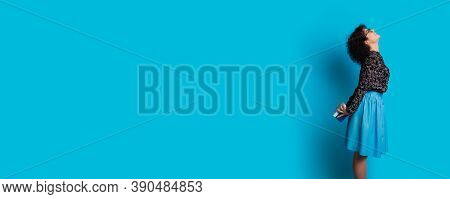 Curly Haired Woman Is Holding A Present At Her Back Smiling On A Blue Wall With Free Space In A Dres
