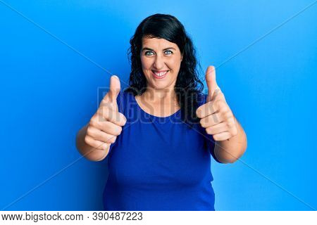 Plus size brunette woman wearing casual blue shirt approving doing positive gesture with hand, thumbs up smiling and happy for success. winner gesture. 