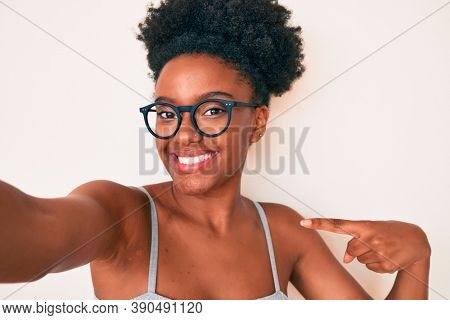 Young african american woman wearing casual clothes and glasses making selfie by the camera pointing finger to one self smiling happy and proud 
