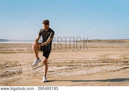 Motivated focused young sportsman exercising at the beach, stetching