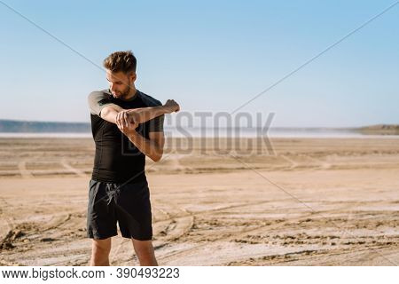 Motivated focused young sportsman exercising at the beach, stetching