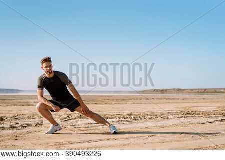Motivated focused young sportsman exercising at the beach, stetching