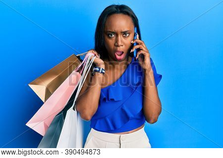Young african woman holding shopping bags and speaking on smartphone in shock face, looking skeptical and sarcastic, surprised with open mouth 