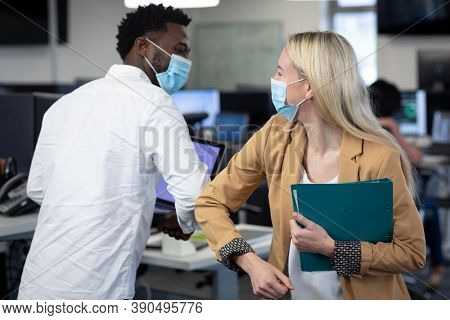 African American man and Caucasian woman in a modern office wearing face masks greeting each other by touching elbows. Hygiene and social distancing in workplace during Coronavirus Covid 19 pandemic.