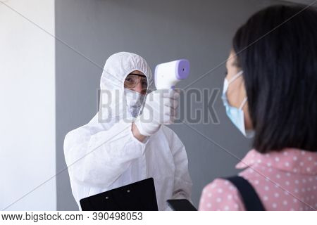Mixed race businesswoman having temperature taken by worker in protective suit before being allowed in to office. Hygiene and social distancing in workplace during Coronavirus Covid 19 pandemic.