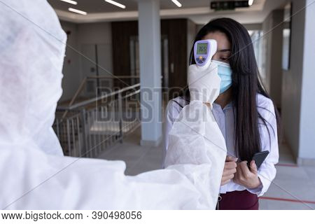 Mixed race businesswoman having temperature taken by worker in protective suit before being allowed in to office. Hygiene and social distancing in workplace during Coronavirus Covid 19 pandemic.