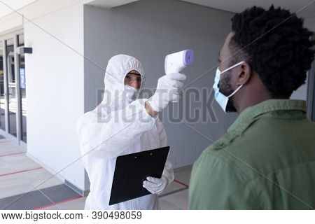 African American businessman having temperature taken by worker in protective suit before being allowed in to office. Hygiene and social distancing in workplace during Coronavirus Covid 19 pandemic.