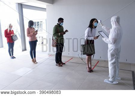 Multi ethnic group of male and female coworkers wearing face masks have temperature checked before entering office. Hygiene and social distancing in workplace during Coronavirus Covid 19 pandemic.