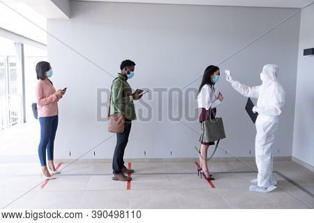 Multi ethnic group of male and female coworkers wearing face masks have temperature checked before entering office. Hygiene and social distancing in workplace during Coronavirus Covid 19 pandemic.