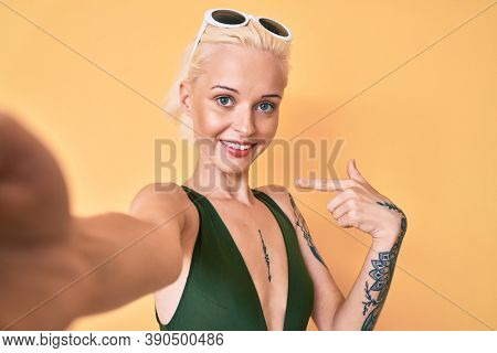 Young blonde woman with tattoo wearing swimwear taking a selfie pointing finger to one self smiling happy and proud 