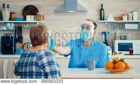 Doctor With Face Mask Checking Senior Woman Temperature Using Gun Thermometer During Home Visit. Soc