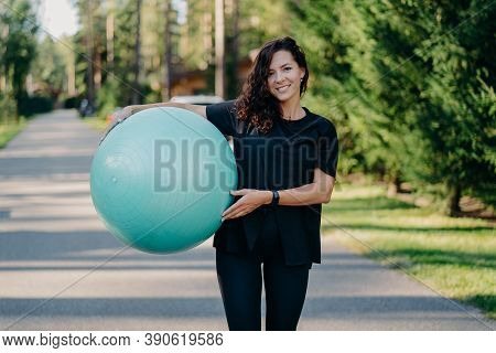 Outdoor Shot Of Brunette Woman Poses With Big Fitball, Dressed In Sportclothes, Has Training In Open