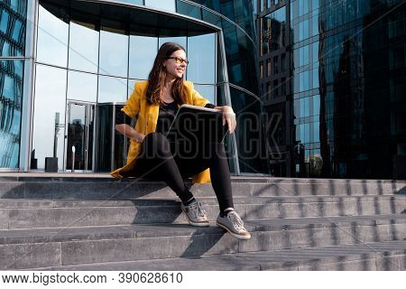 Freelancer With A Laptop Sat On The Steps Near The Business Center, A Young Woman In A Yellow Jacket