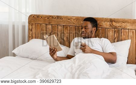 Morning Read. Black Millennial Guy Reading Book Drinking Coffee Lying In Comfortable Bed In Bedroom 