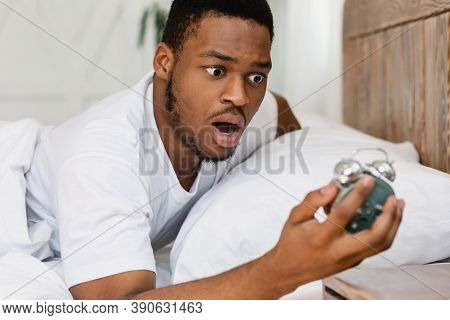 Oversleeping Concept. Shocked Black Guy Looking At Broken Alarm Clock Waking Up Late For Work In The