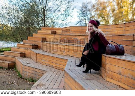 A Joyful Girl Sits On A Summer Theater Bench, Wooden In A Burgundy Coat And Biret, In The Fall Again