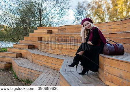 A Cheerful Lady Sits On A Summer Theater Bench, Made Of Wood In A Burgundy Coat And Biret, An Adult 