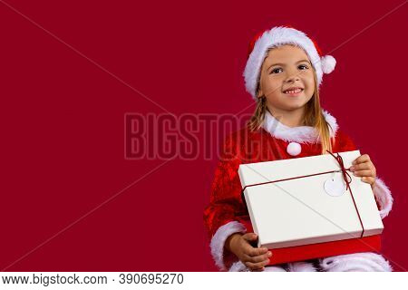 Portrait Of A Blondine Little Girl In Santa Claus Dress. The Girl Looks At The Camera Smiling And Op