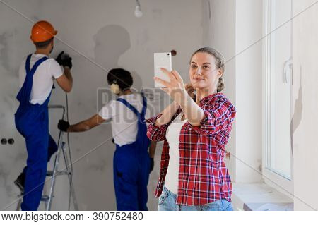 Young Woman Smiles And Takes A Selfie On The Phone. In The Background, Handymen In Overalls Are Dril
