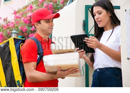 Serious Client Holding Tablet And Reading Order Details. Professional Deliveryman In Red Cap And Shi