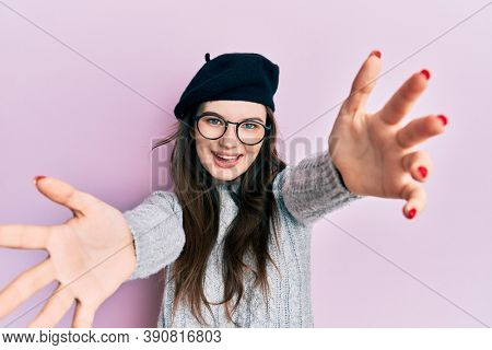 Young beautiful caucasian girl wearing french look with beret looking at the camera smiling with open arms for hug. cheerful expression embracing happiness. 
