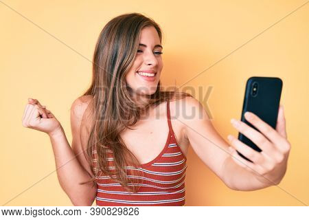 Beautiful young caucasian woman taking a selfie photo with smartphone screaming proud, celebrating victory and success very excited with raised arm 