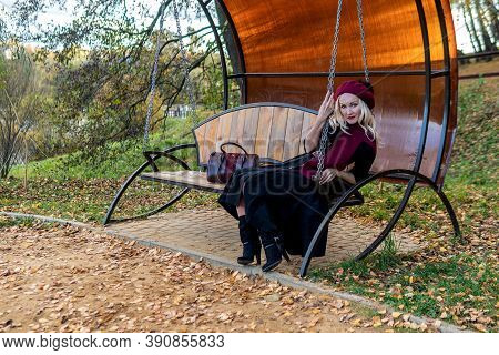 A Happy Girl Sits On A Garden Swing, Forged In A Burgundy Coat And Biret, Smiles At The Camera, In T