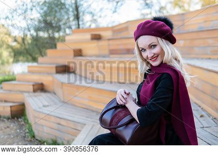 A Smiling Woman Sits On A Summer Theater Bench, Made Of Wood In A Burgundy Coat And Biret, An Adult 