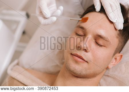 Young Attractive Man Relaxing At Spa Center While Beautician Applying Peeling Lotion On His Forehead