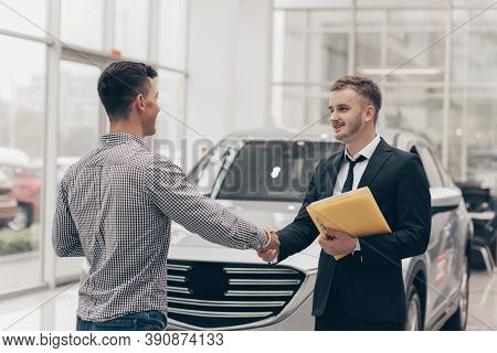 Professional Car Salesman Smiling, Shaking Hands With His Male Customer In Front Of A New Automobile