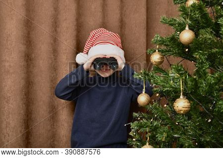 The Boy Smiles And Watches Gifts Near The Christmas Tree, Looks Out Through Binoculars. Dressed In A