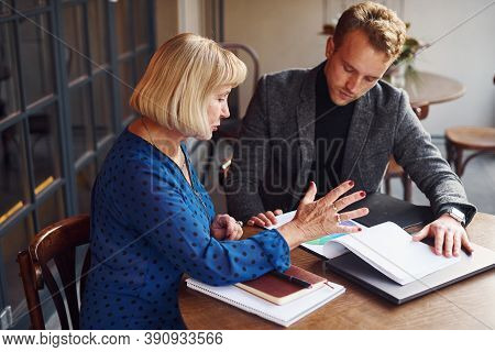Working With Documents. Young Guy In Formal Clothes Have A Business Talk With Old Woman In Cafe.