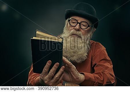 A wise old man with a long gray beard in a bowler hat and glasses is reading an old book. Black background. 