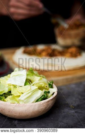 closeup of a bowl with chopped lettuce on a table, and a young man in the background preparing a durum or a burrito, with chicken meat cooked with different vegetables, such as onion or red pepper