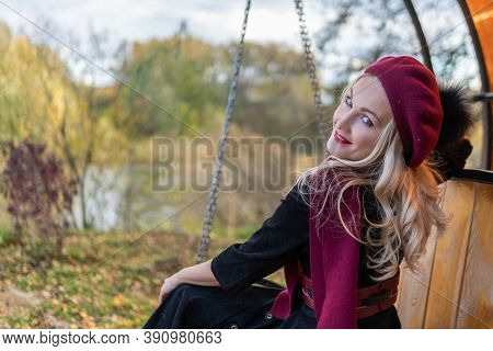 A Cheerful Lady Sits On A Garden Swing, Forged In A Burgundy Coat And Biret, An Adult With Tender Li