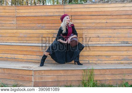 Happy Lady Sits On A Bench Of A Summer Theater, Wooden In A Burgundy Coat And Biret, Looking At The 
