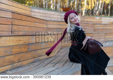 A Happy Girl Sits On A Summer Theater Bench, Wooden In A Burgundy Coat And Biret, Smiles At The Came