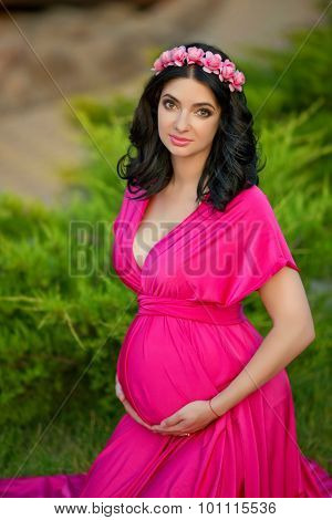 Beautiful Pregnant Woman With Chaplet Of Flowers Wearing In Pink Dress Relaxing In The Park.