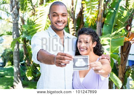 Portrait of happy man with pregnant wife holding sonogram against white background
