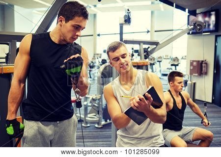 sport, fitness, lifestyle and people concept - men with clipboard taking notes and exercising on gym machine