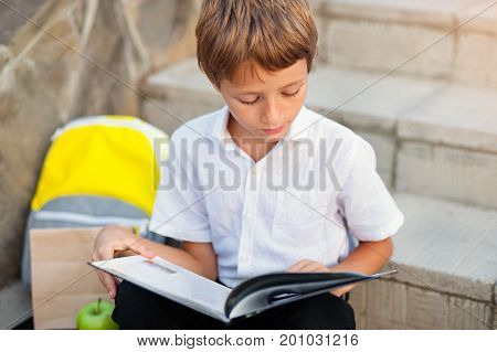 Cute schoolboy studying outdoors on bright autumn day. Boy is reading book with backpack. Back to school. Young student doing homework. Education for small kids.