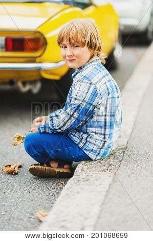 Outdoor fashion portrait of cute little 6 year old boy wearing blue plaid casual shirt and trousers sitting on a sidewalk next to retro yellow car