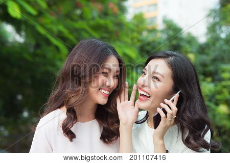 Young asian woman whispering secret into a friends ear while shes on call