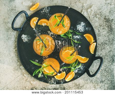 Refreshing cold alcoholic summer citrus cocktail with orange, peppermint and crushed ice in stemless glasses on dark tray over grey concrete background, top view